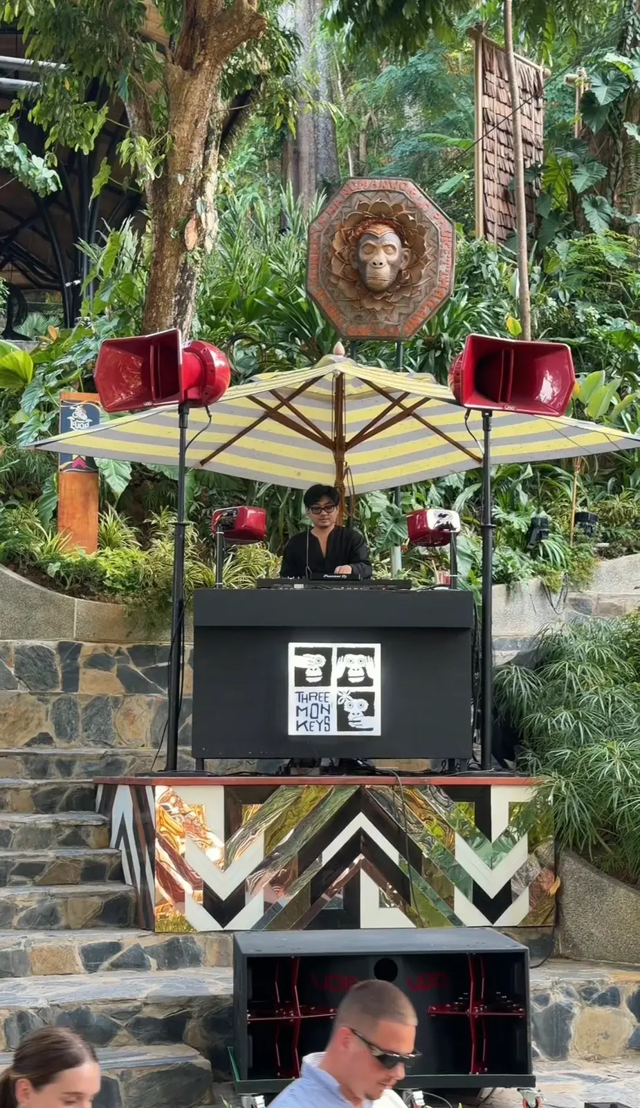 DJ booth beneath an umbrella beside stone steps and tropical plants